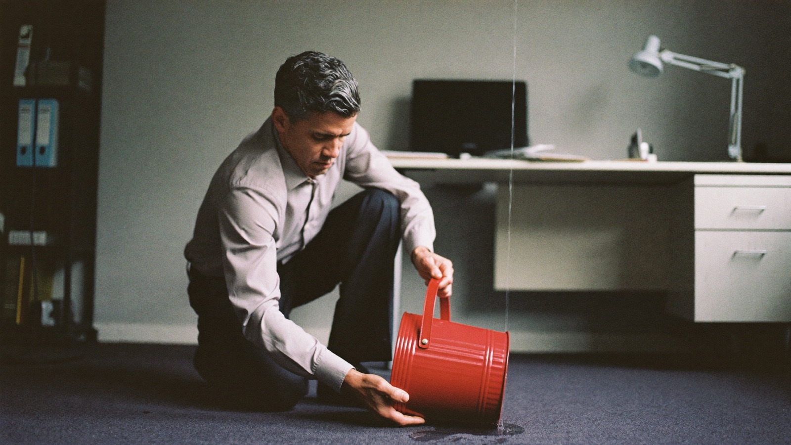 A business owner kneeling in his office, placing a red bucket under a thin water leak.